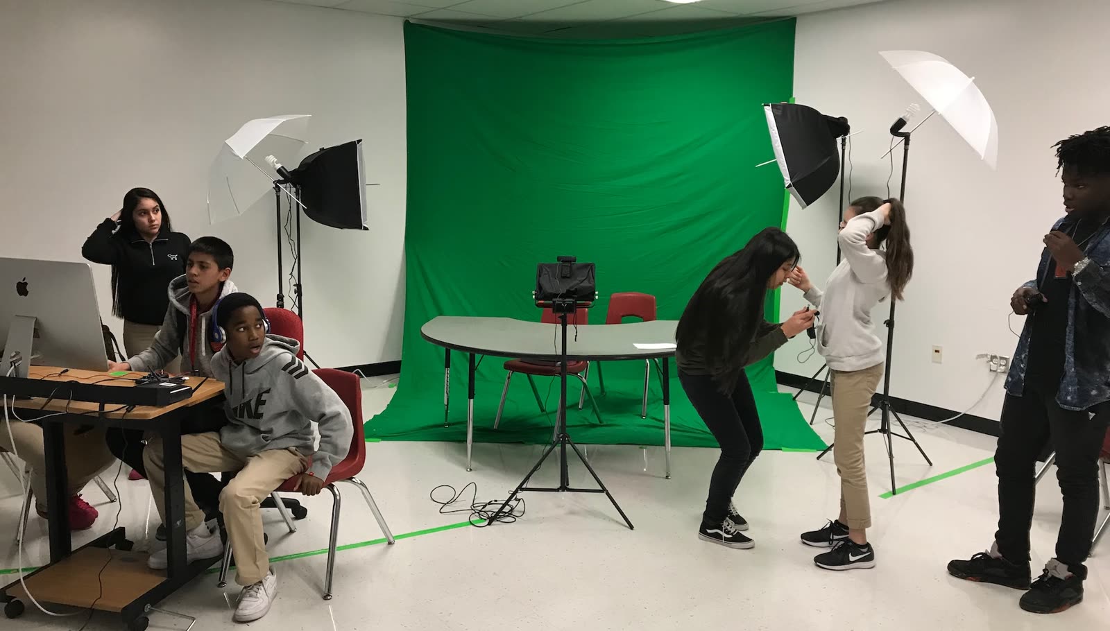 Students at a classroom news studio: one crew at the control desk with a Mac and audio mixer, others setting up the shot in front of a green screen
