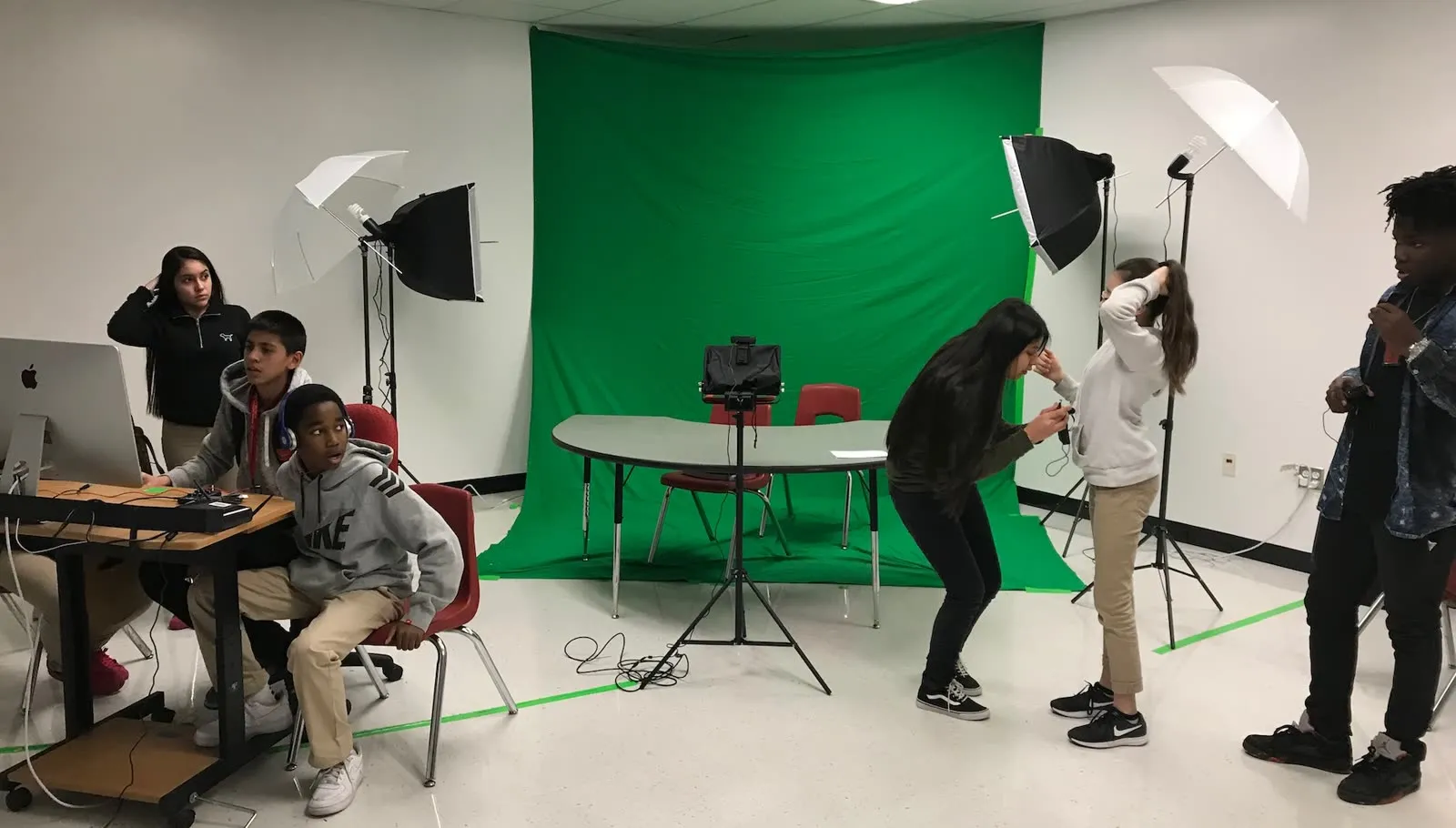 Students at a classroom news studio: one crew at the control desk with a Mac and audio mixer, others setting up the shot in front of a green screen